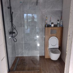Paving House - Niedokończona łazienka z kabiną prysznicową typu walk-in z czarną armaturą, szarymi płytkami imitującymi marmur, drewnopodobną zabudową WC i narzędziami do silikonowania na podłodze.