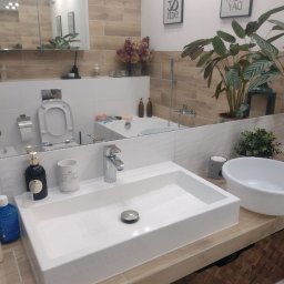 Paving House - Nowoczesna łazienka z prostokątną białą umywalką nablatową, baterią chromowaną, lustrem odbijającym wannę i dekoracje, drewnopodobne płytki na ścianach i wiklinowy kosz pod umywalką.