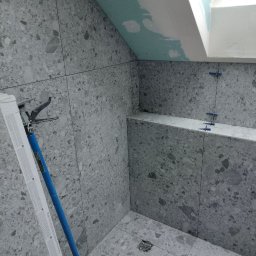 Paving House - Szara łazienka w trakcie wykańczania z widocznymi krzyżykami dystansowymi, drabina oparta o ścianę i surowy sufit z oknem dachowym.