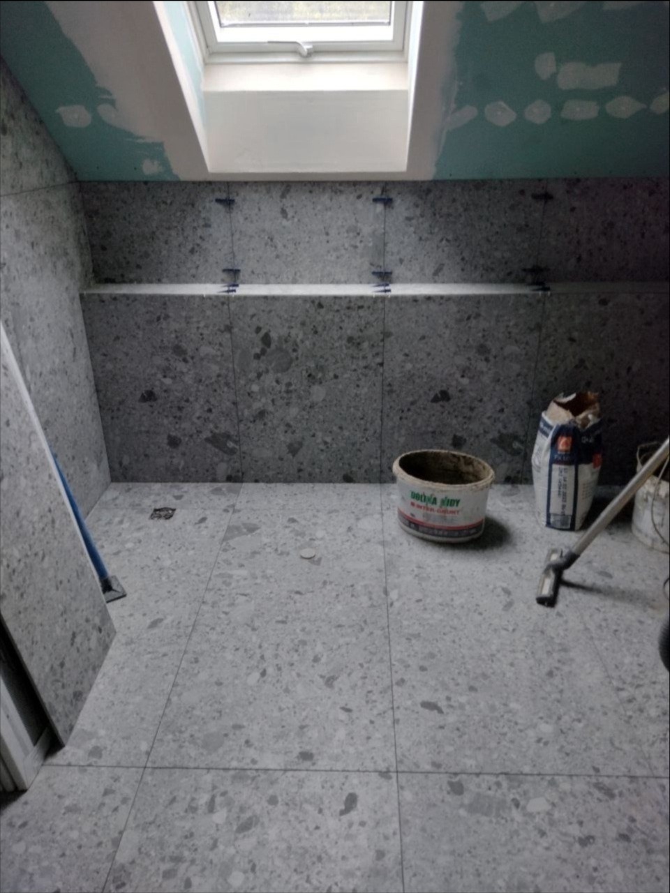 Łazienka w trakcie wykańczania: szare płytki imitujące lastryko na podłodze i ścianach, widoczne krzyżyki dystansowe, wiadro z zaprawą i okno dachowe.