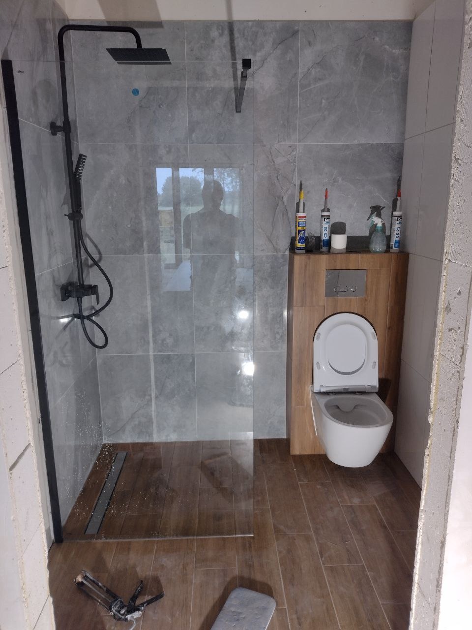 Niedokończona łazienka z kabiną prysznicową typu walk-in z czarną armaturą, szarymi płytkami imitującymi marmur, drewnopodobną zabudową WC i narzędziami do silikonowania na podłodze.