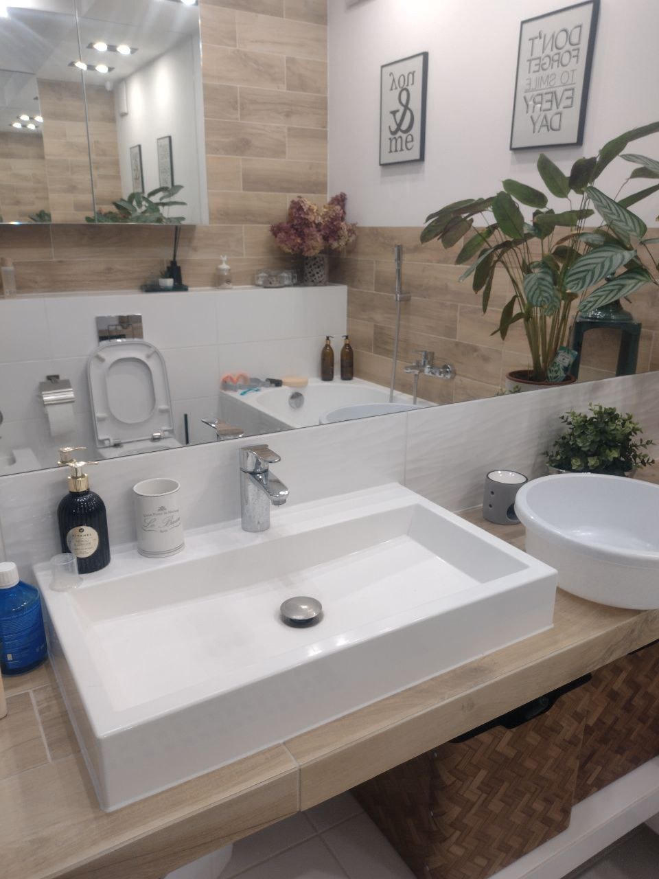 Nowoczesna łazienka z prostokątną białą umywalką nablatową, baterią chromowaną, lustrem odbijającym wannę i dekoracje, drewnopodobne płytki na ścianach i wiklinowy kosz pod umywalką.