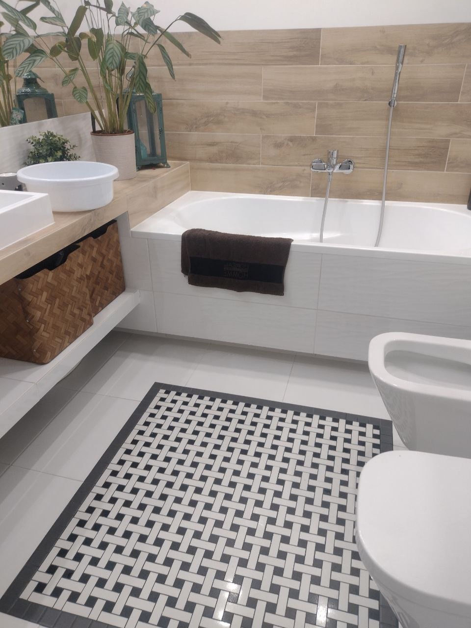 Nowoczesna łazienka z drewnopodobnymi płytkami na ścianie, białą wanną z brązowym ręcznikiem, umywalką z wiklinowymi koszami, toaletą i czarno-białą mozaiką na podłodze.