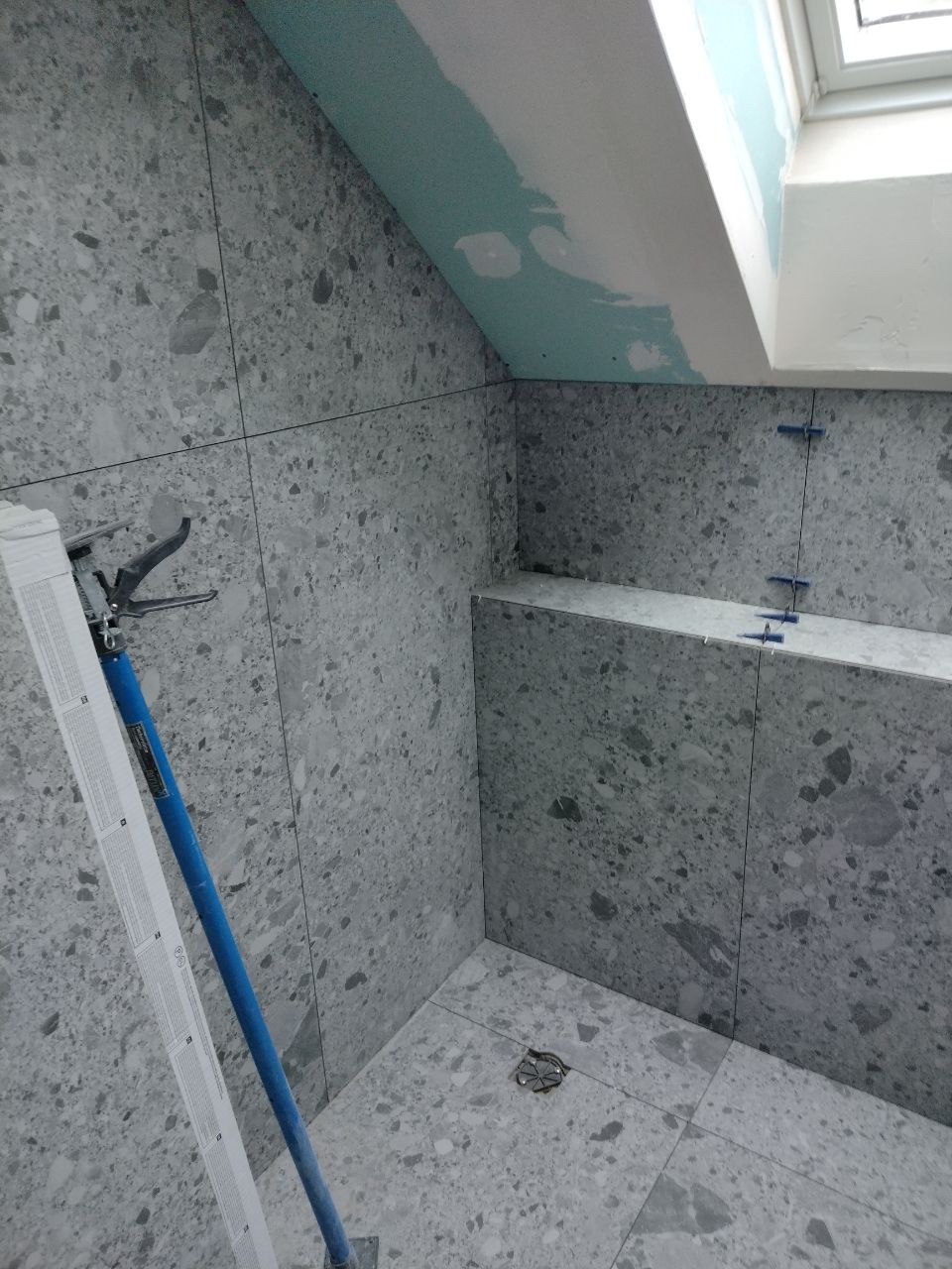 Szara łazienka w trakcie wykańczania z widocznymi krzyżykami dystansowymi, drabina oparta o ścianę i surowy sufit z oknem dachowym.