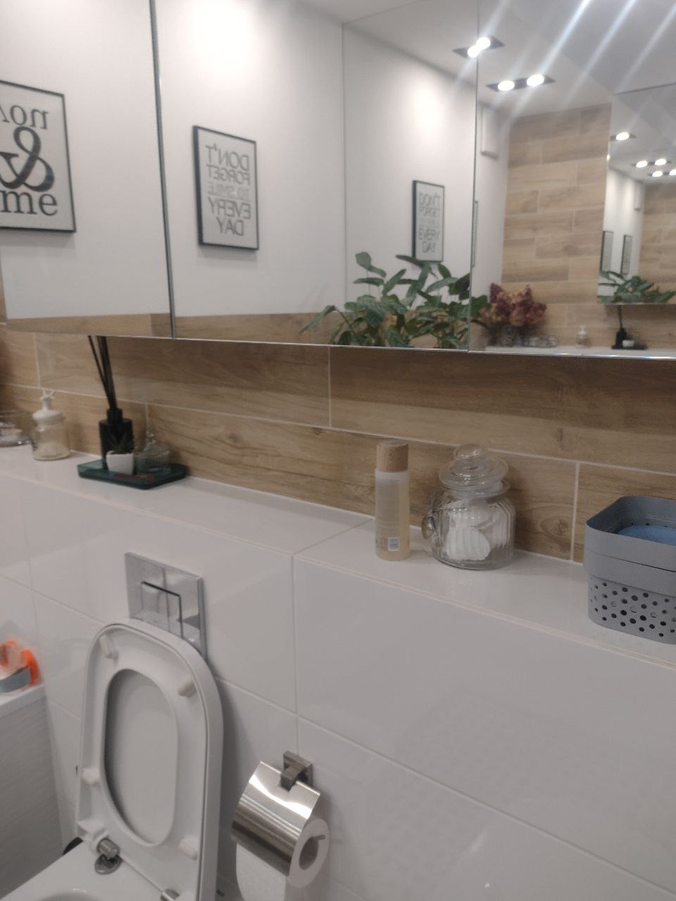 Jasna łazienka z białymi płytkami i drewnopodobnym pasem płytek nad umywalką, otwarty sedes, dekoracje na półce pod lustrem, rośliny.