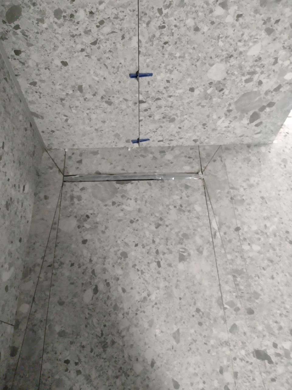 Fragment łazienki w trakcie układania szarych płytek lastryko, widoczne krzyżyki dystansowe i folia zabezpieczająca odpływ liniowy.