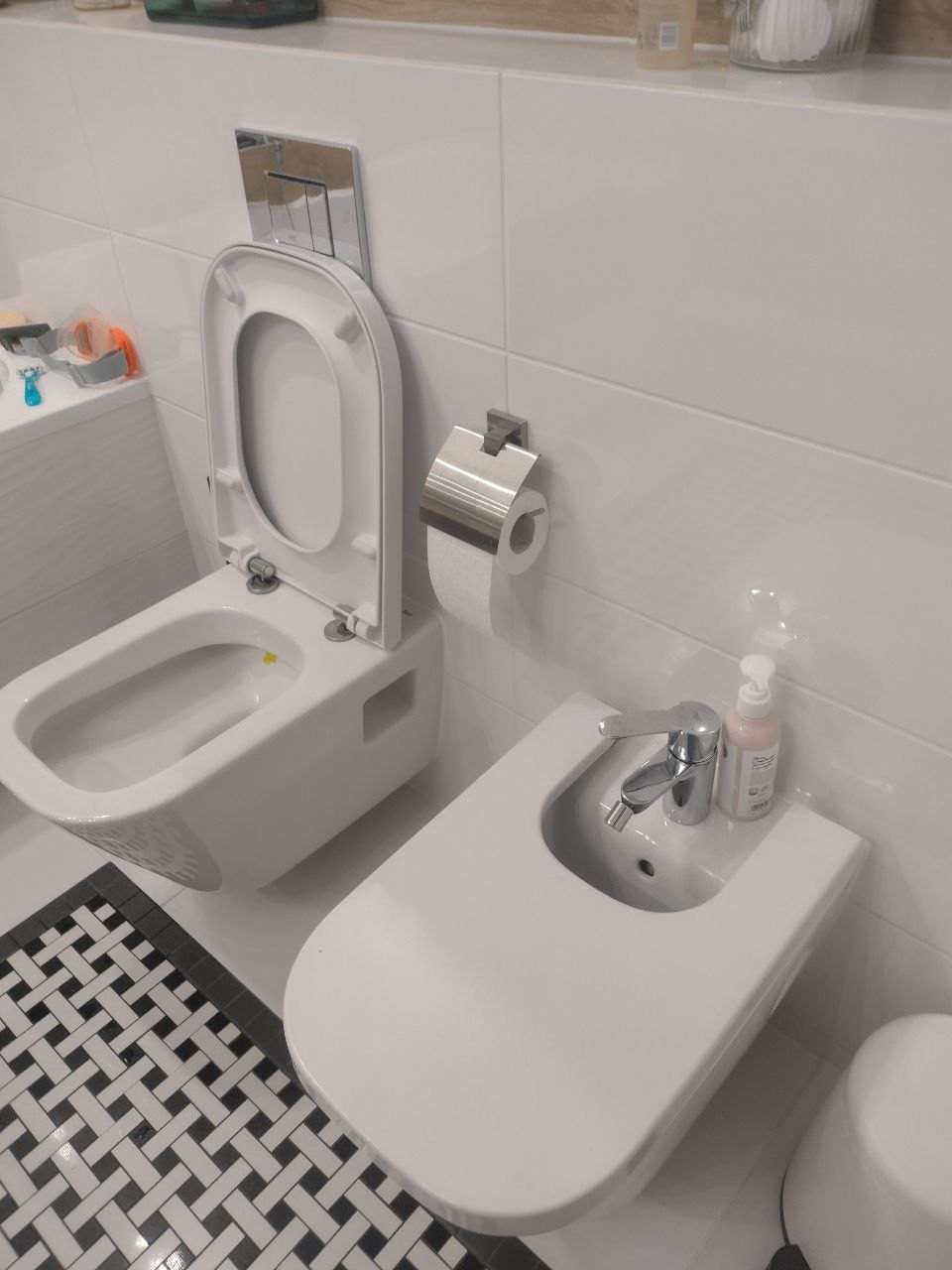 Nowoczesna, biała łazienka z podwieszaną toaletą z otwartą deską i bidetem, białe płytki na ścianach, czarno-biała mozaika na podłodze, minimalistyczny styl.