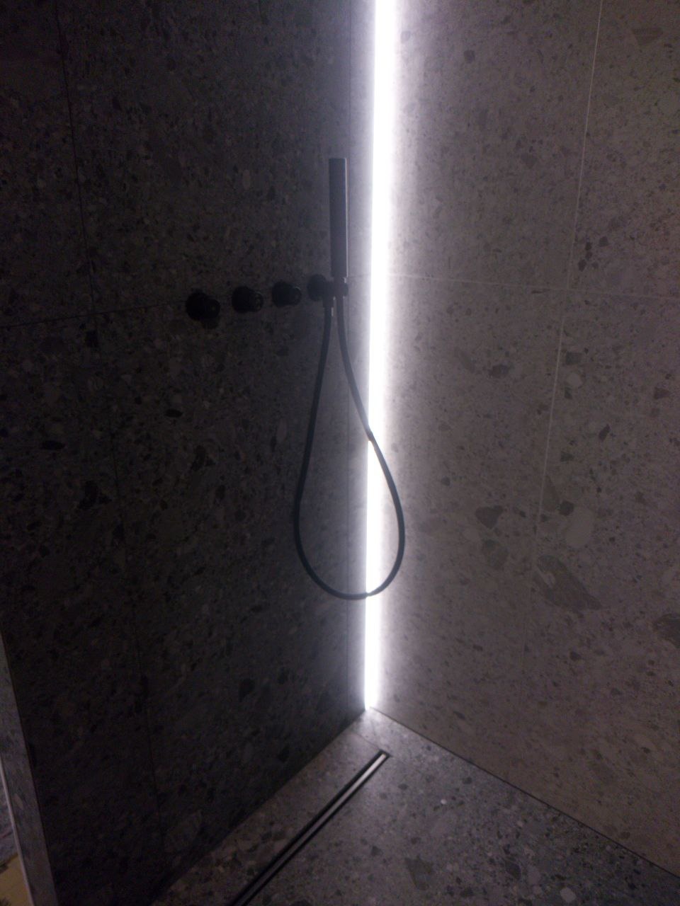 Nowoczesna kabina prysznicowa z szarej terazzo, podświetlana pionową listwą LED, czarna armatura i odpływ liniowy.