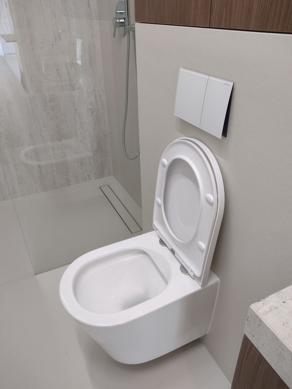 Nowoczesna, podwieszana toaleta z deską wolnoopadającą, zamontowana w minimalistycznej łazience z kabiną prysznicową typu walk-in i szarymi płytkami.