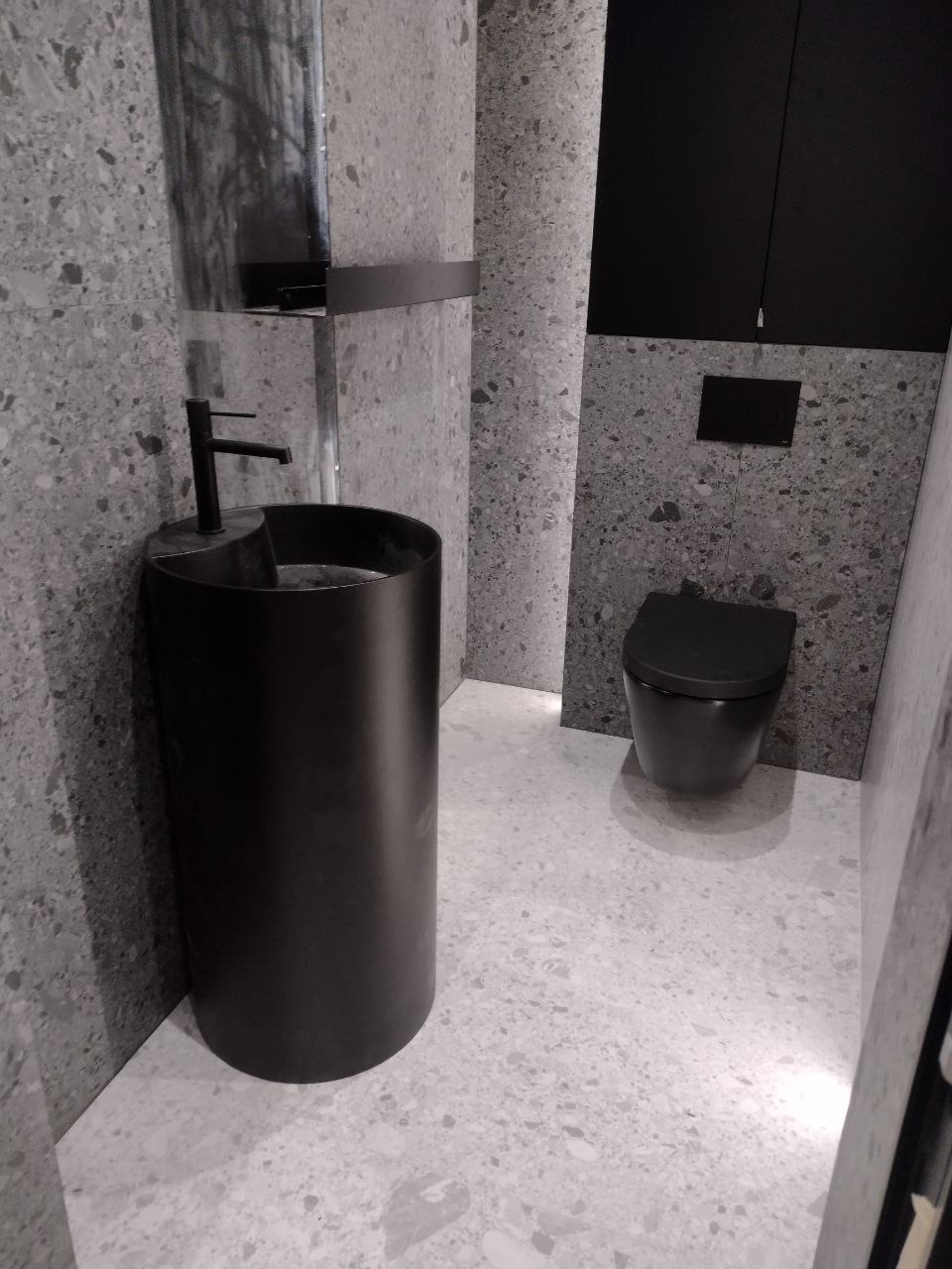 Nowoczesna łazienka w szarościach z czarną, cylindryczną umywalką wolnostojącą i podwieszaną toaletą. Ściany i podłoga wyłożone płytkami imitującymi lastryko.