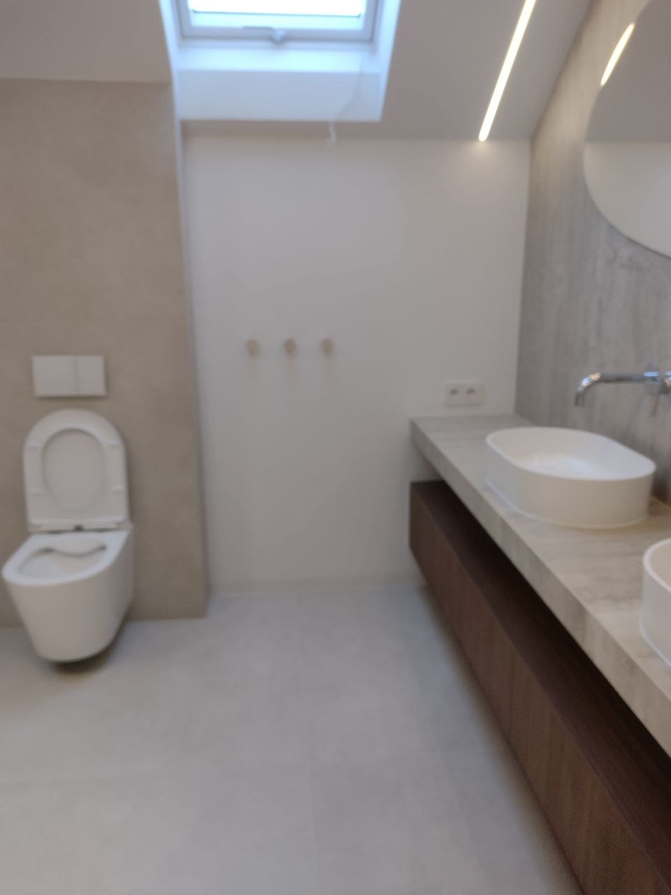 Nowoczesna łazienka z toaletą wiszącą, umywalką nablatową na drewnianej szafce i oknem dachowym, oświetlona listwą LED.