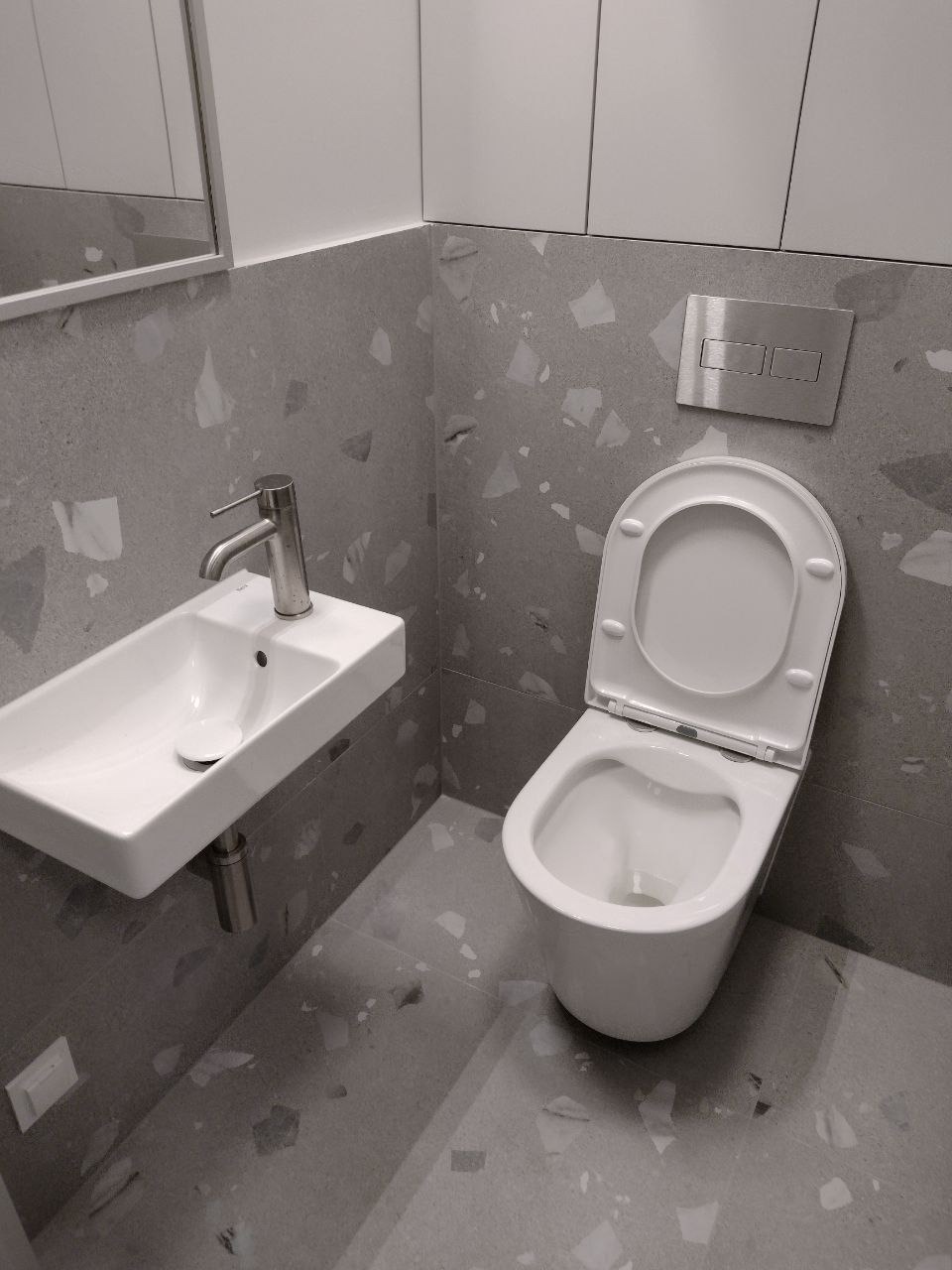 Nowoczesna łazienka w szarościach: podwieszana umywalka z chromowaną baterią, toaleta z podtynkowym systemem spłukiwania, płytki imitujące lastryko na ścianach i podłodze, minimalistyczne szafki...