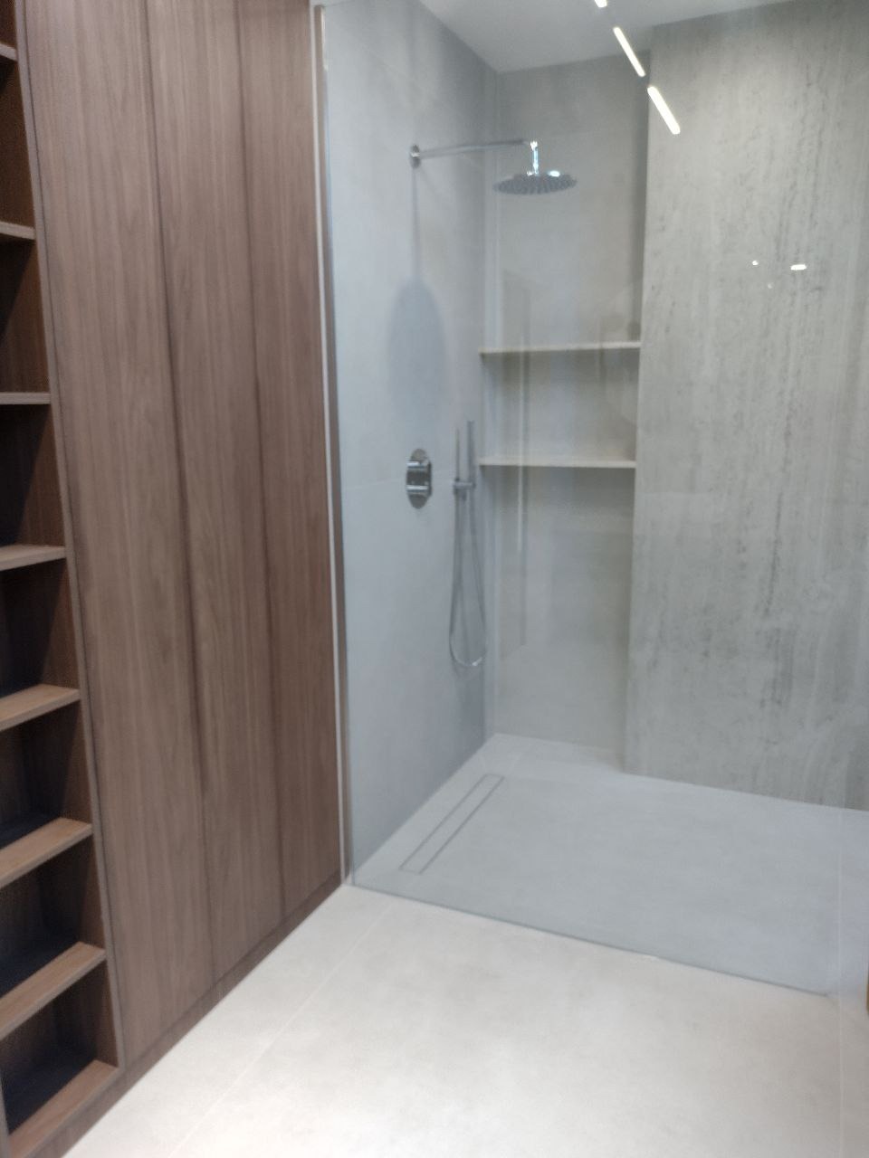 Nowoczesna kabina prysznicowa walk-in z odpływem liniowym, szklaną ścianką i półkami wewnątrz, sąsiadująca z zabudową meblową z drewnopodobnymi frontami.