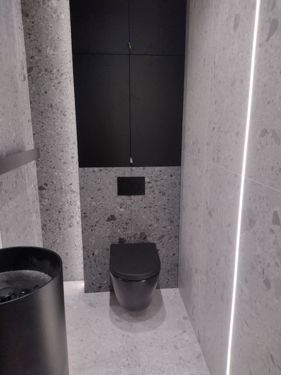 Nowoczesna toaleta w szaro-czarnej kolorystyce z umywalką na blacie i podświetleniem LED w pionowej szczelinie między płytkami.