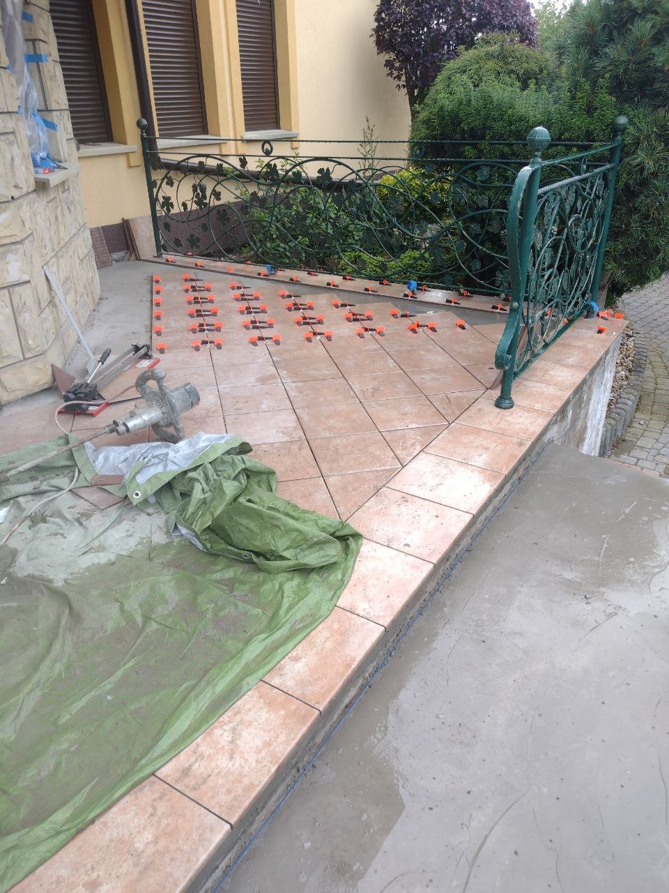 Układanie terakoty na schodach wejściowych, widoczne narzędzia i system poziomowania płytek, przykryte plandeką.