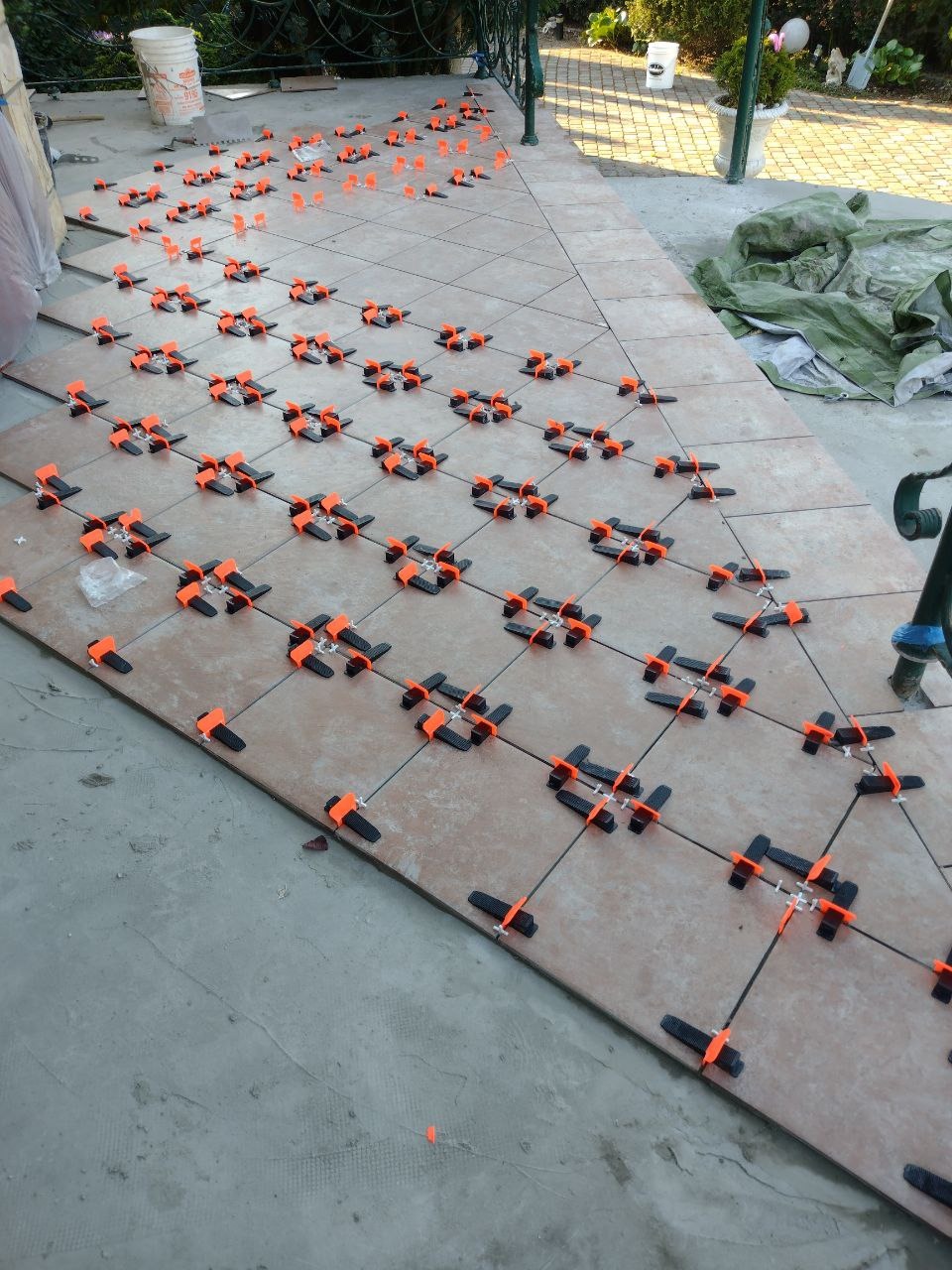 Układanie brązowych płytek na zewnątrz z użyciem pomarańczowo-czarnych klipsów poziomujących, widoczne wiadro i plandeka w tle.