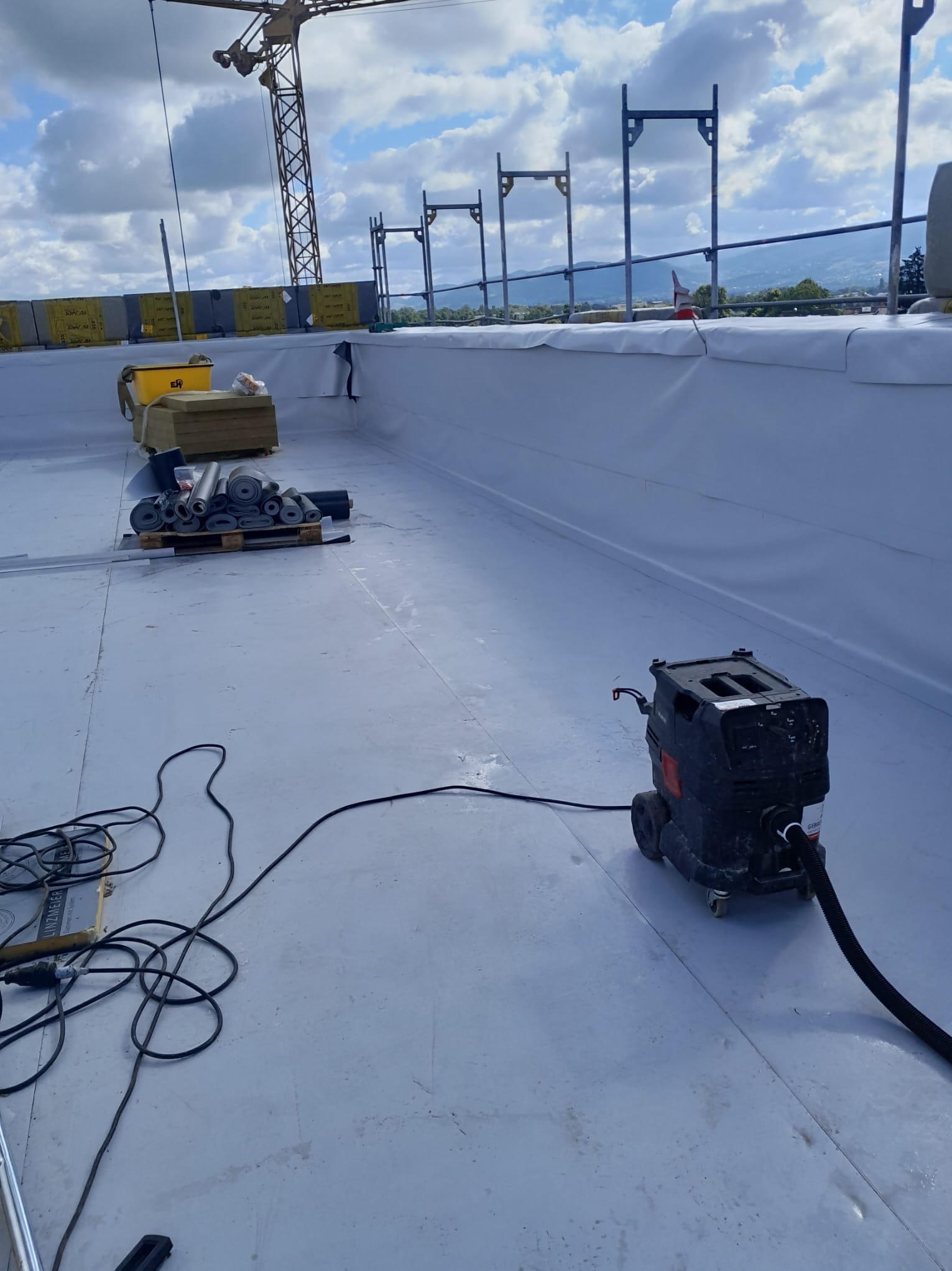 Biała membrana dachowa na płaskim dachu z widocznymi rolkami materiału izolacyjnego, narzędziami i maszyną czyszczącą, w tle dźwig budowlany i krajobraz.