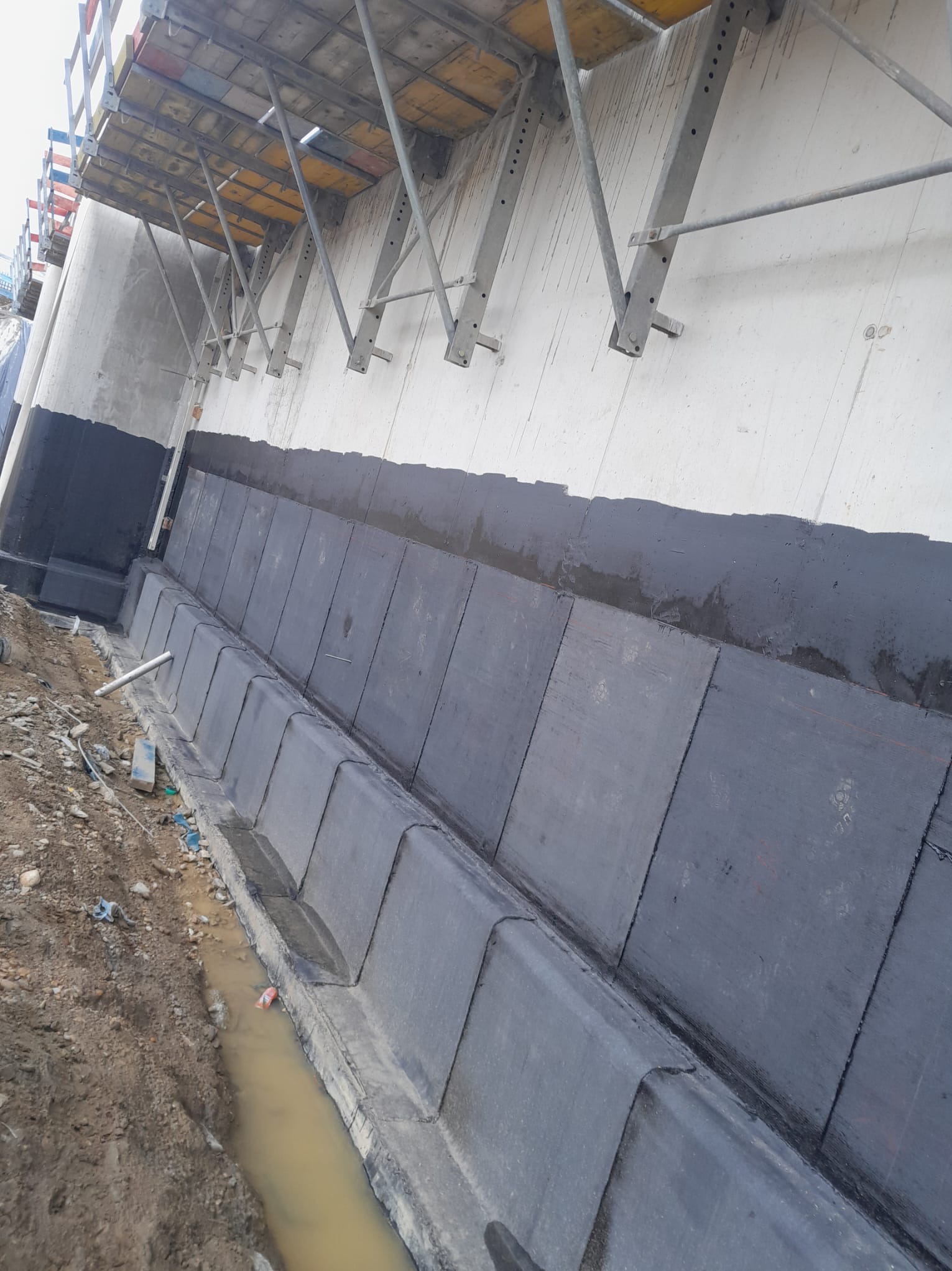 Fundament budynku w trakcie izolacji bitumicznej, z widocznymi panelami izolacyjnymi i konstrukcją wsporczą szalunku.