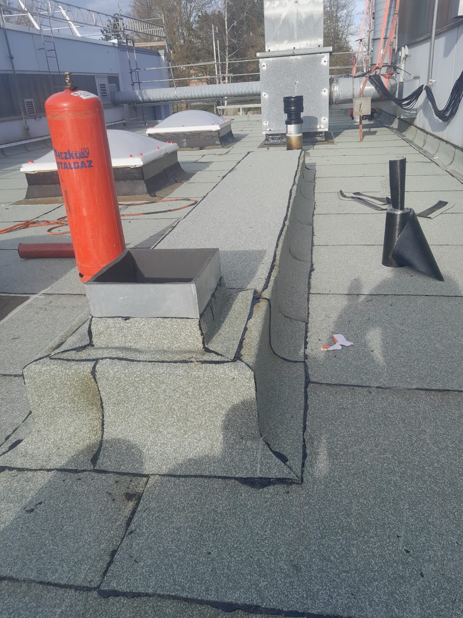 Płaski dach pokryty papą termozgrzewalną, widoczny komin z obróbką blacharską, butla gazowa Totalgaz, świetliki dachowe oraz narzędzia dekarskie, fragment orzeźwiającego nieba w tle.