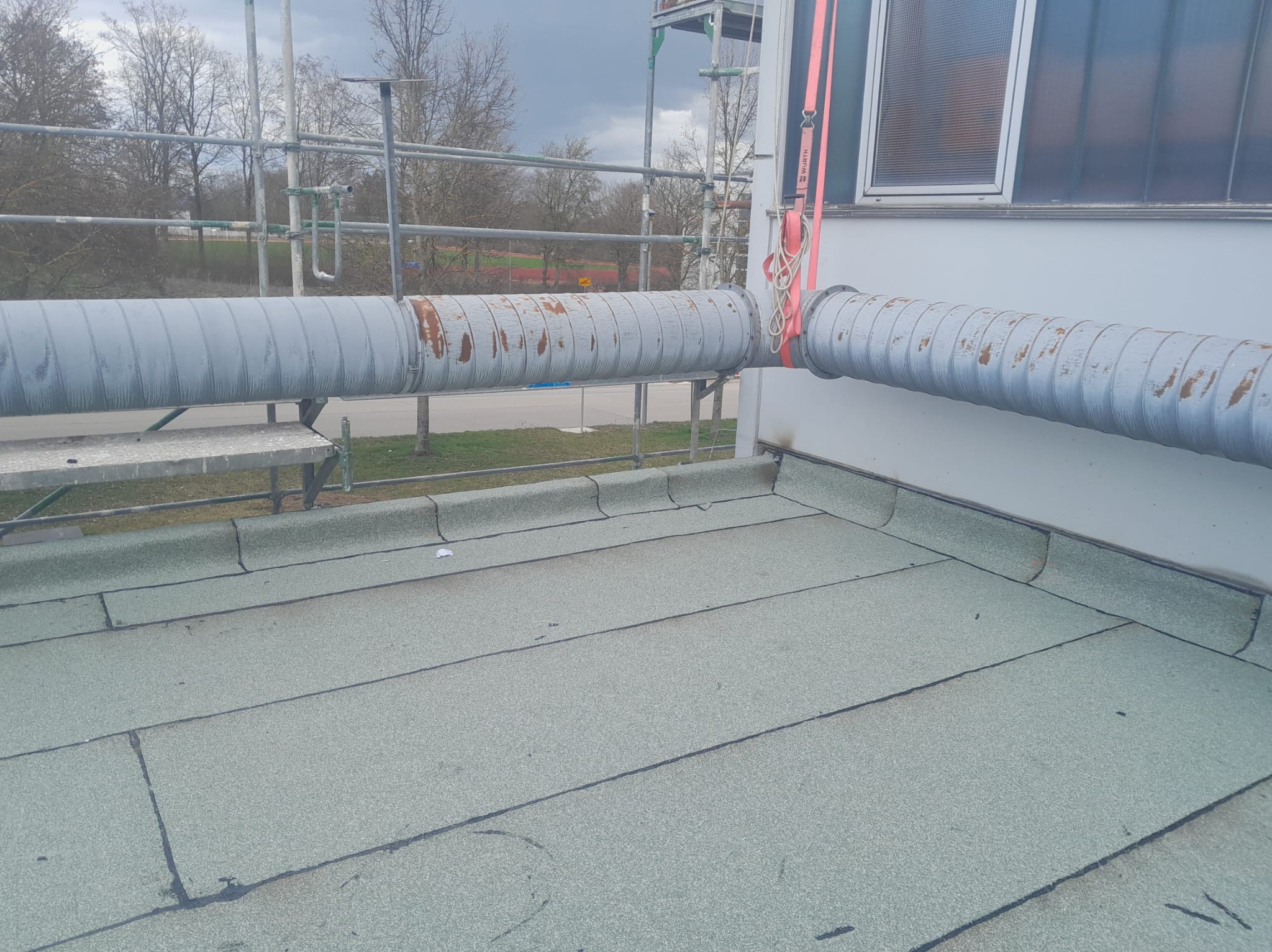 Szary dach pokryty papą z widocznymi rurami wentylacyjnymi i rusztowaniem w tle.