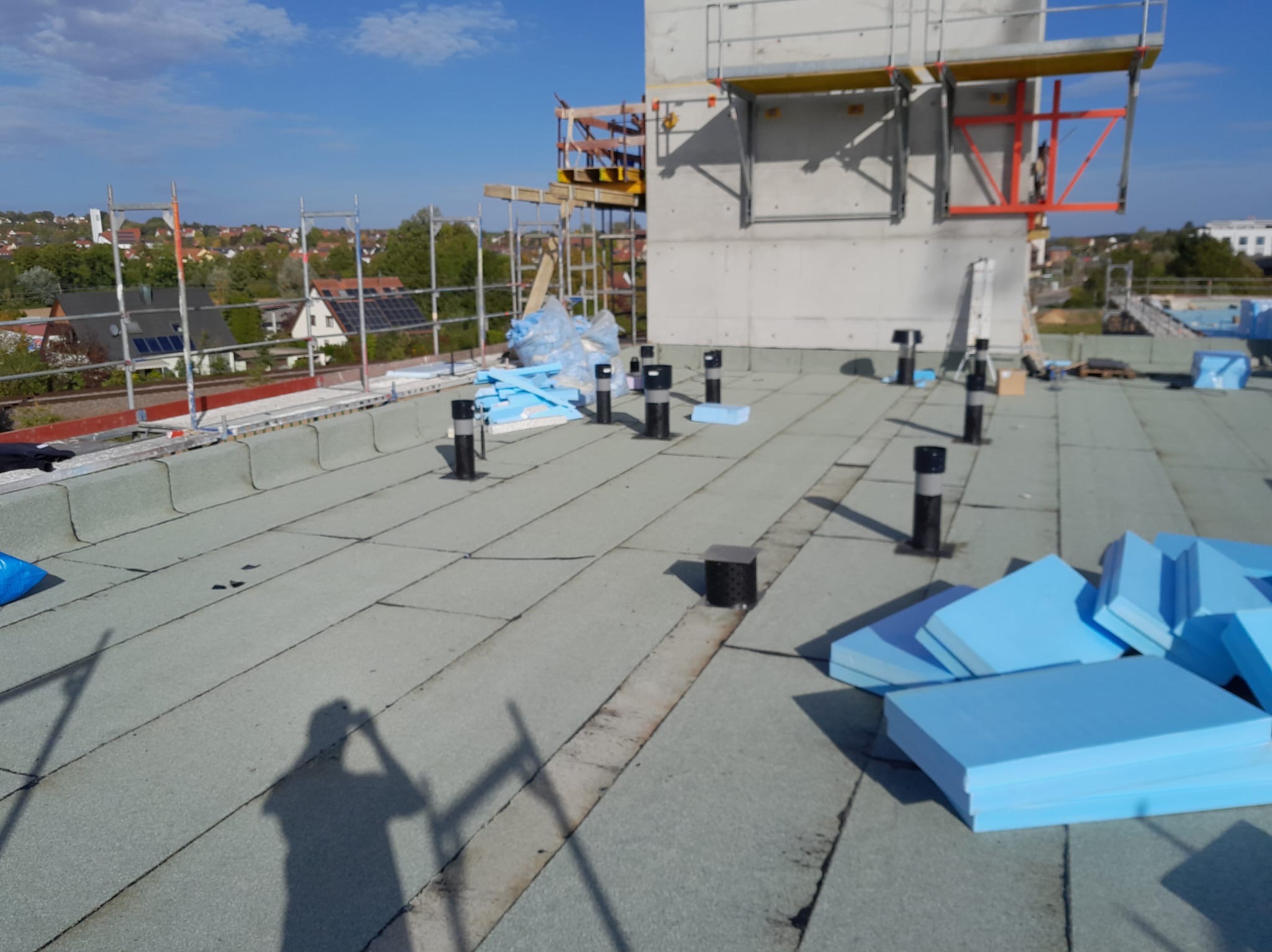 Przygotowanie do izolacji dachu płaskiego: widoczne czarne kominki wentylacyjne, niebieskie płyty izolacyjne i cień osoby na pokryciu dachowym, w tle budynek w trakcie budowy i panorama miasta.