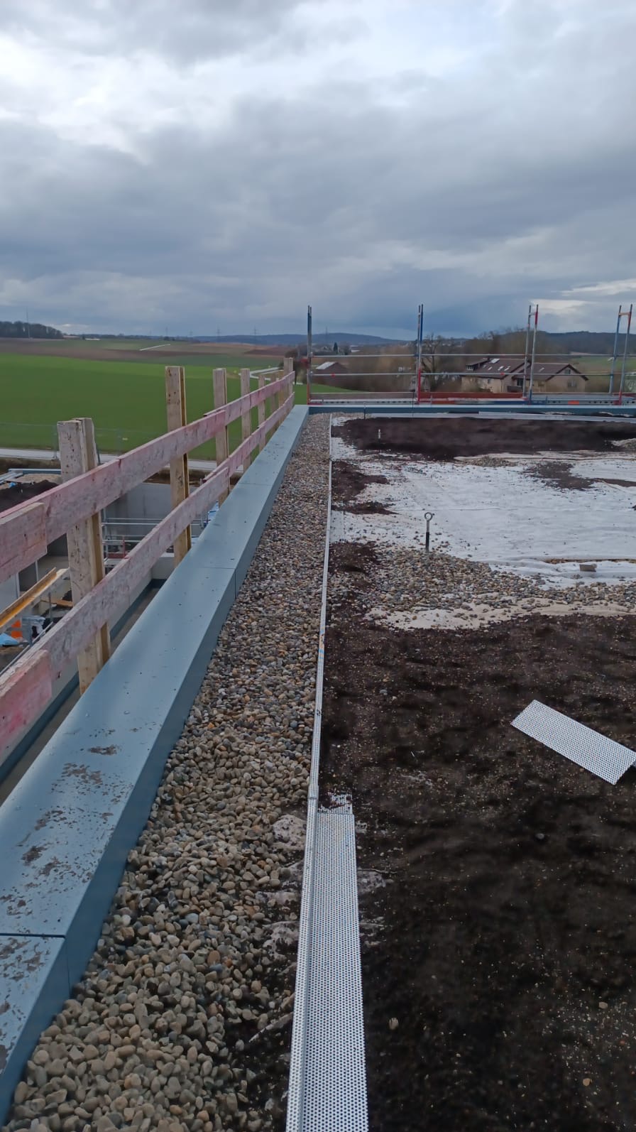 Płaski dach w trakcie budowy z widoczną barierką ochronną, warstwą drenażową z kamyków i elementami wentylacji, na tle pochmurnego nieba i zielonych pól.