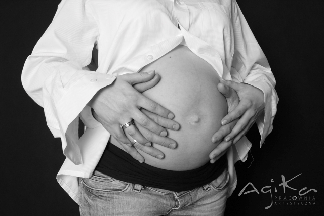 Czarno-białe zdjęcie ciążowego brzuszka w dłoniach kobiety z pierścionkami i mężczyzny, ubranych w białą koszulę i jeansy, na ciemnym tle.