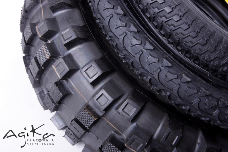 Dwa czarne opony rowerowe z agresywnym bieżnikiem, ułożone jedna na drugiej, z widocznym logo 'Agika Pracownia Artystyczna' w lewym dolnym rogu.