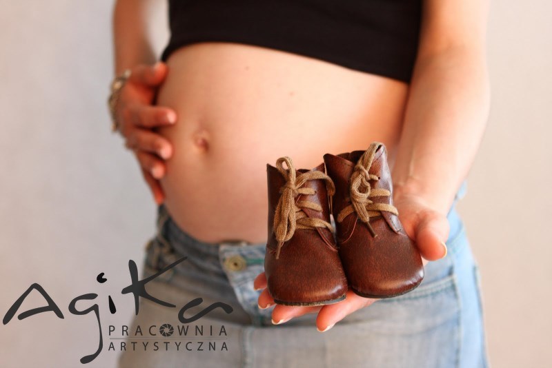 Ciężarna kobieta trzymająca w dłoni parę małych, brązowych skórzanych bucików ze sznurowadłami, z widocznym brzuchem i logo fotografa w lewym dolnym rogu.