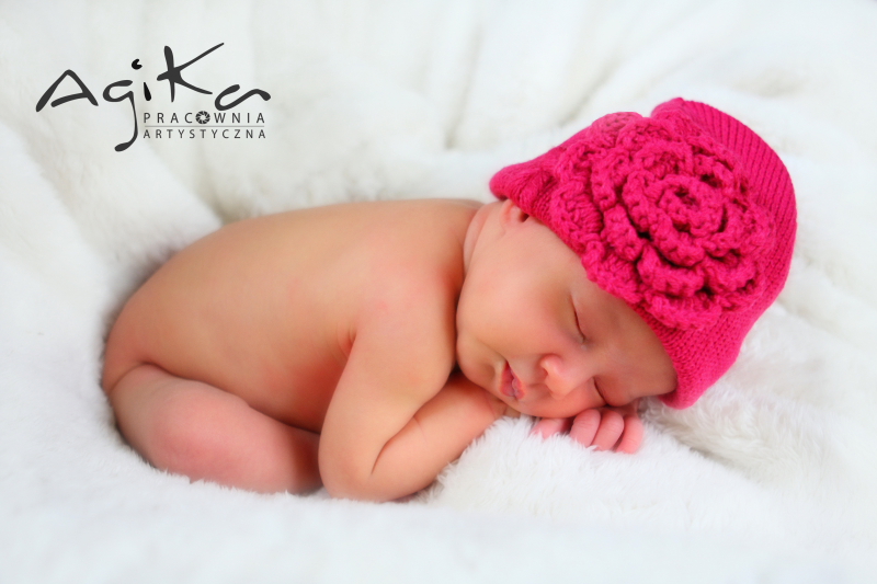 Śpiące niemowlę w różowej, dzierganej czapeczce z kwiatem na miękkim, białym futerku.