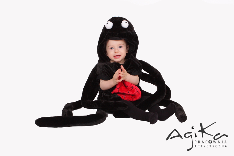 Uśmiechnięte niemowlę w czarnym kostiumie ośmiornicy z dużymi oczami i czerwonym elementem na brzuchu, siedzące na białym tle.