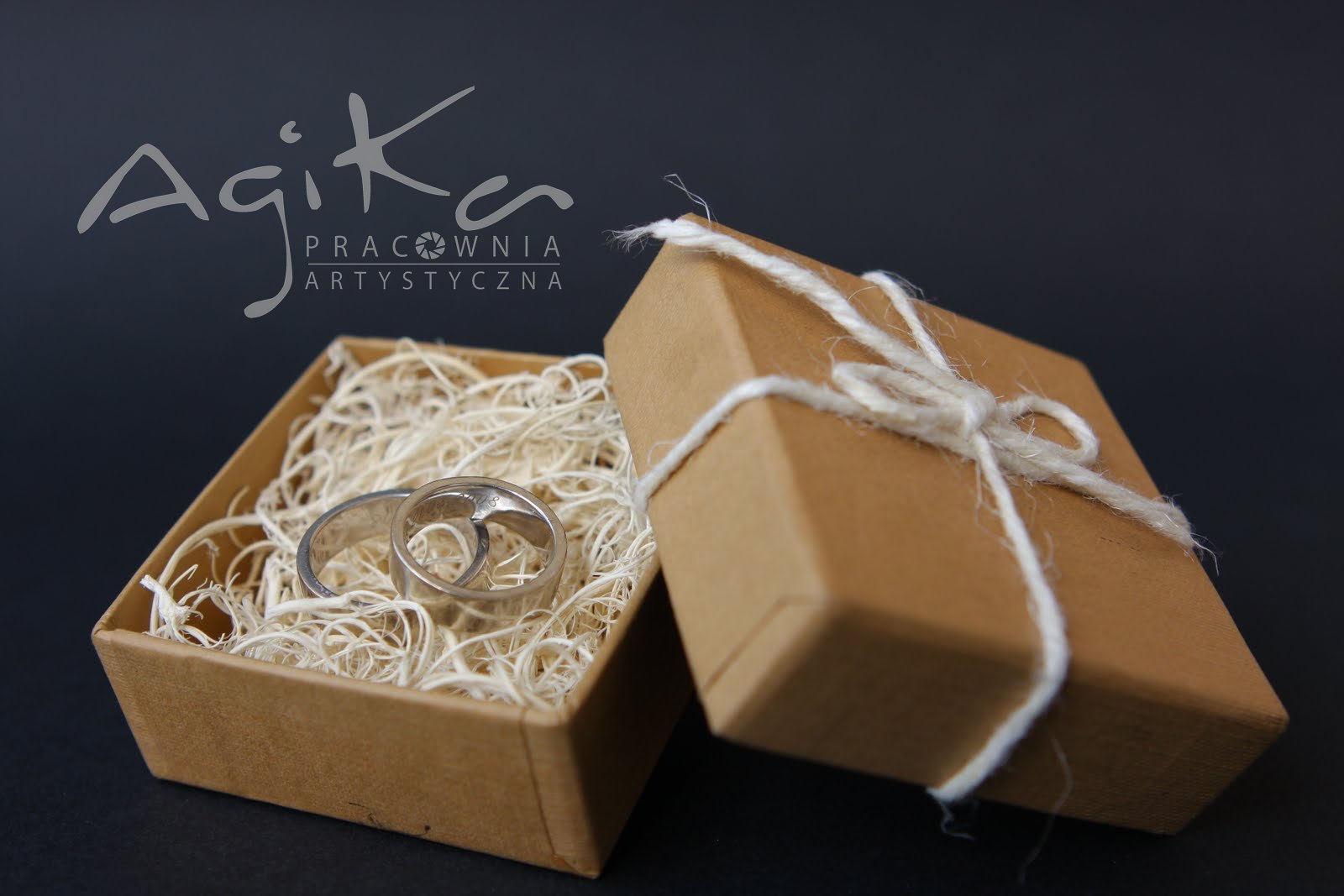 Dwa srebrne obrączki w brązowym pudełku wypełnionym siankiem, obok pudełko z zawiązanym sznurkiem, na ciemnym tle, widoczne logo firmy Agika Pracownia Artystyczna.