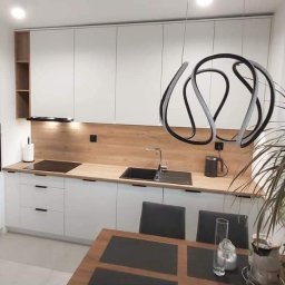 Meble wroclaw - Nowoczesna kuchnia z białymi frontami i drewnianym blatem, czarny zlewozmywak i płyta indukcyjna, nad blatem wisząca lampa o abstrakcyjnym kształcie, w tle stół z krzesłami i roślina doniczkowa.