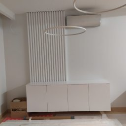 Meble wroclaw - Biała szafka RTV z lamelami ściennymi w tle, oświetlona okrągłą lampą sufitową we Wrocławiu, widoczne materiały montażowe.