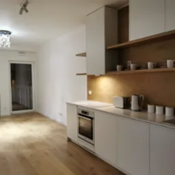 Nowoczesna kuchnia w minimalistycznym stylu z białymi frontami szafek i drewnianym panelem na ścianie, oświetlona punktowymi lampami sufitowymi i podświetleniem blatu, z widocznym sprzętem AGD...