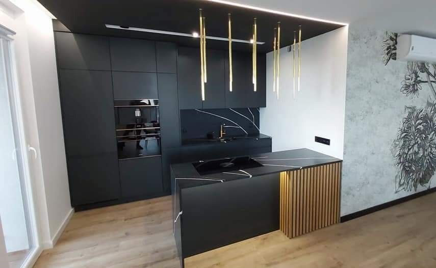 Nowoczesna kuchnia w czerni z wyspą, blatem z czarnego kamienia z białymi żyłami, złotymi lampami wiszącymi i drewnianymi lamelami na wyspie, oświetlona listwą LED pod sufitem.