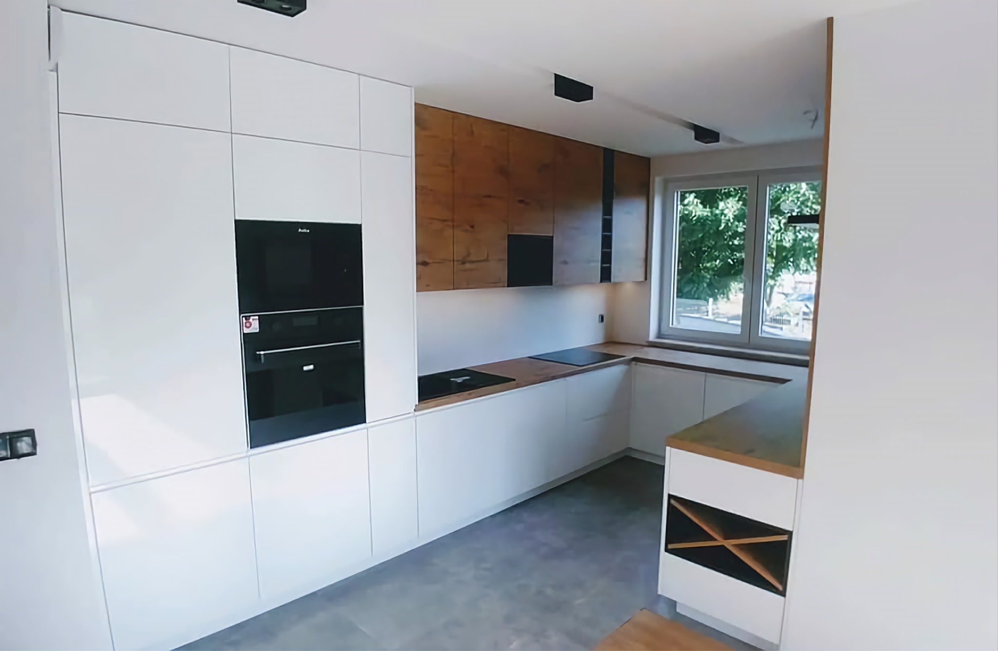 Nowoczesna kuchnia z białymi, matowymi frontami i górnymi szafkami w okleinie drewnopodobnej, czarne wbudowane piekarniki i płyta indukcyjna, szare płytki na podłodze, minimalistyczne oświetlenie...