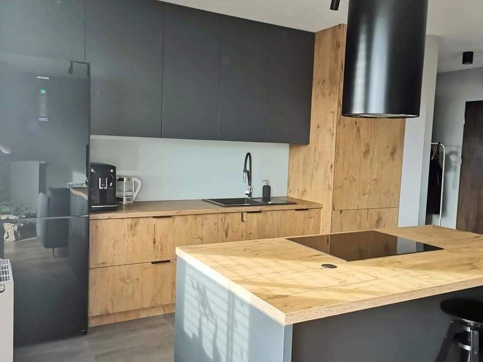 Nowoczesna kuchnia z wyspą, fronty szafek w kolorze grafitowym i drewnopodobnym, czarny okap cylindryczny, czarny sprzęt AGD, szara podłoga