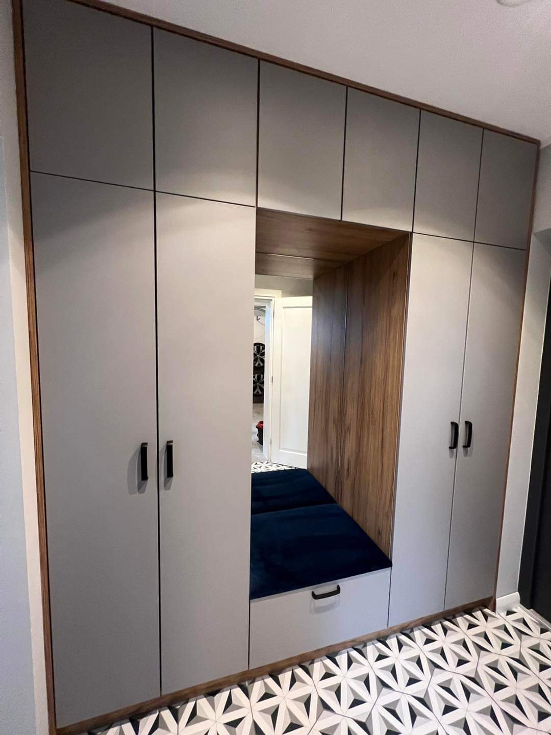 Szara szafa wnękowa z siedziskiem z granatową tapicerką, drewnianym wykończeniem i wzorzystą podłogą.