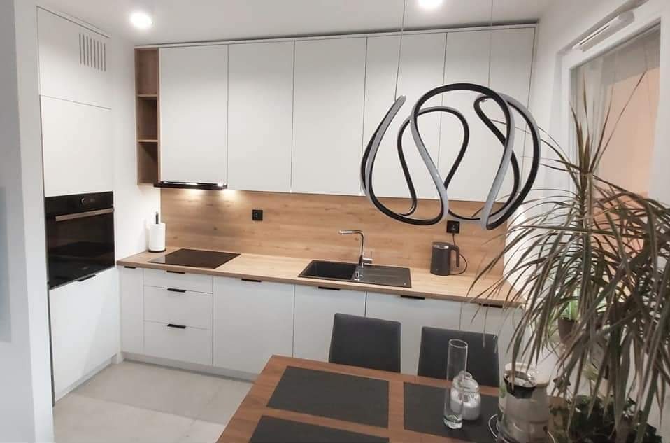 Nowoczesna kuchnia z białymi frontami i drewnianym blatem, czarny zlewozmywak i płyta indukcyjna, nad blatem wisząca lampa o abstrakcyjnym kształcie, w tle stół z krzesłami i roślina doniczkowa.