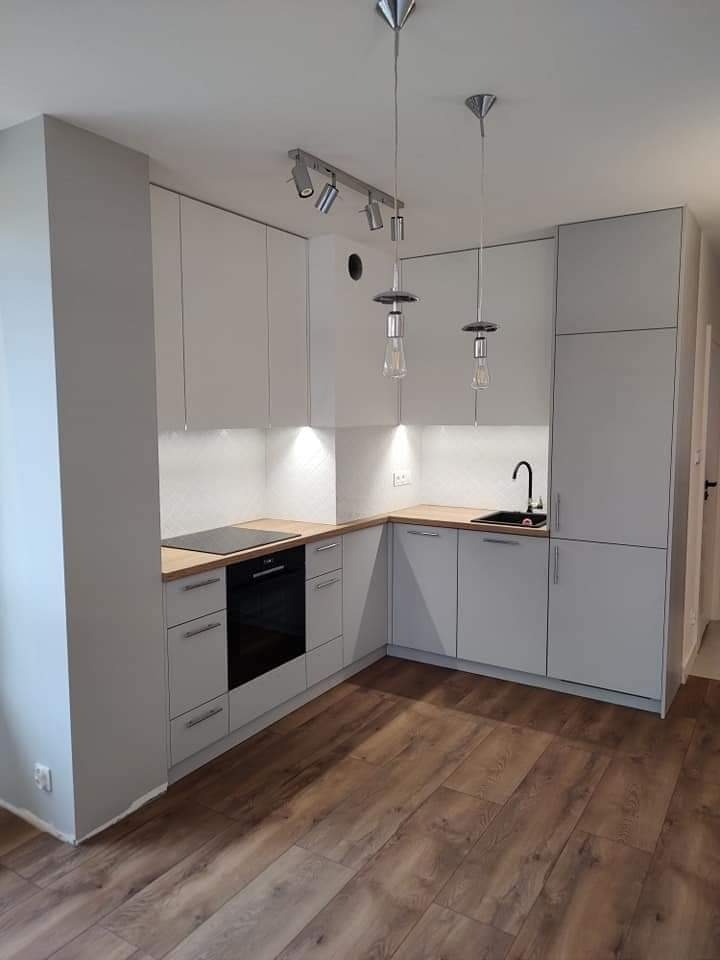 Jasna kuchnia narożna z drewnianym blatem, oświetleniem punktowym i wiszącymi lampami z żarówkami Edisona, na podłodze panele imitujące drewno.