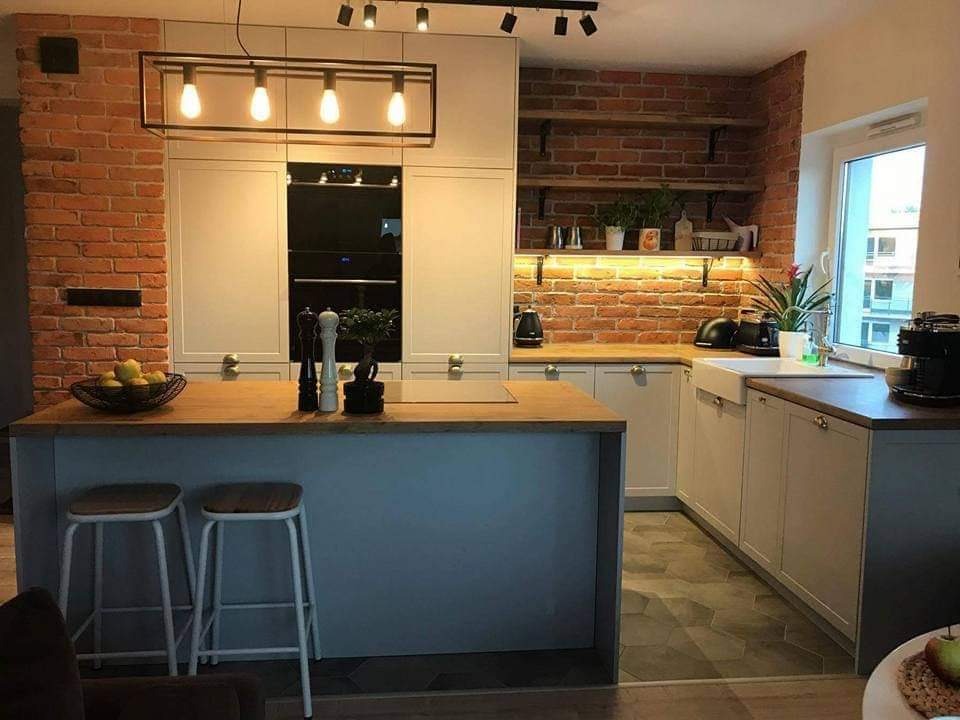 Jasna kuchnia z ceglaną ścianą, drewnianym blatem i wyspą, oświetlona wiszącymi lampami i ledowym paskiem pod półkami.