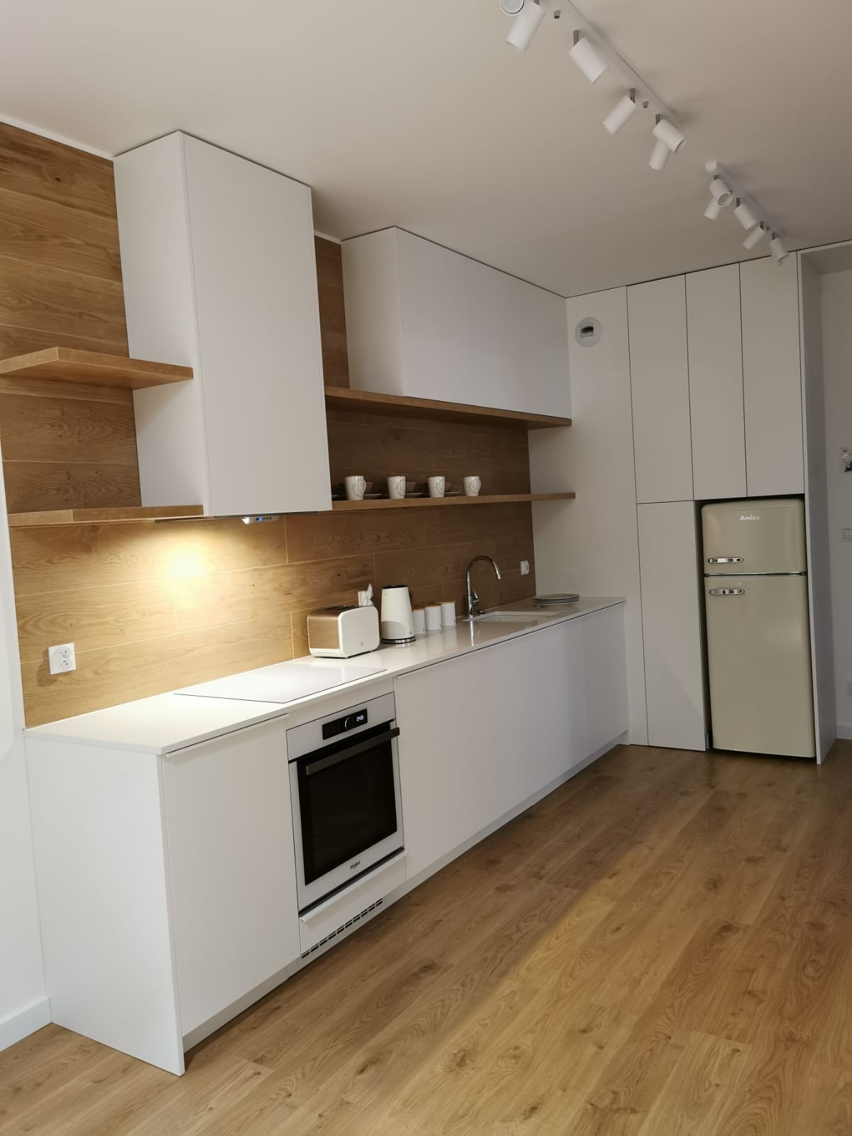 Nowoczesna kuchnia z białymi szafkami, drewnianym panelem ściennym i otwartymi półkami, oświetlona punktowymi lampami sufitowymi, z beżową lodówką w stylu retro.