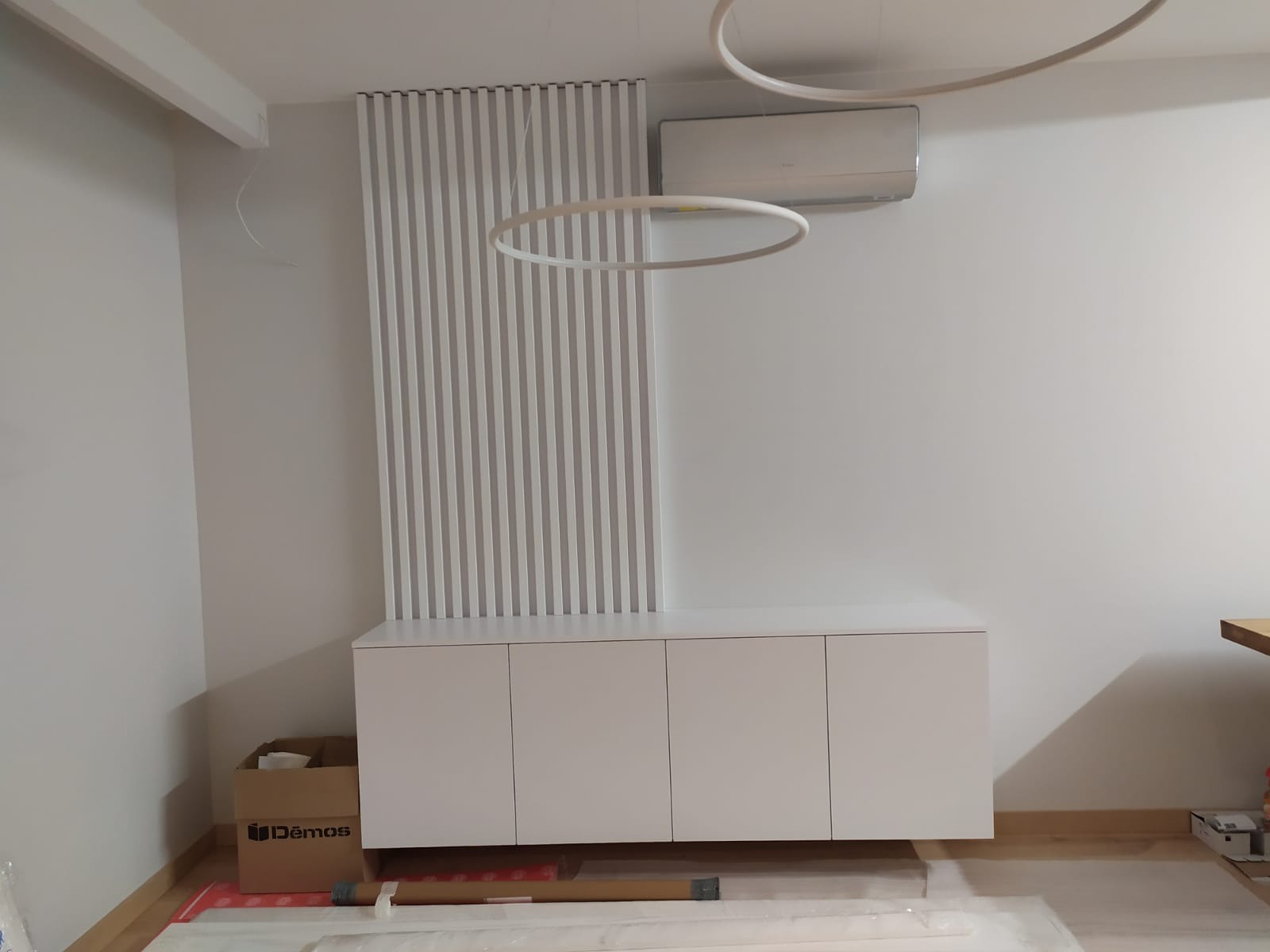 Biała szafka RTV z lamelami ściennymi w tle, oświetlona okrągłą lampą sufitową we Wrocławiu, widoczne materiały montażowe.