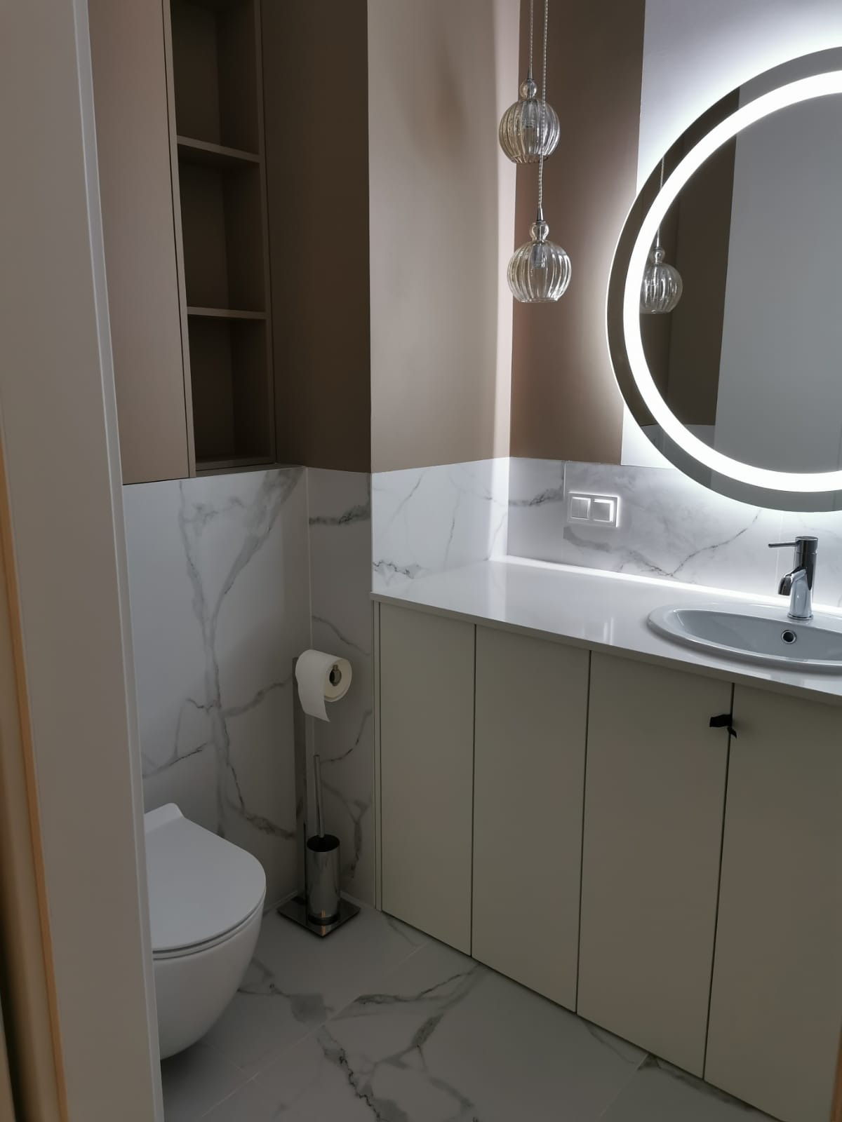 Nowoczesna łazienka z toaletą, szafkami pod umywalką z białym blatem, okrągłym lustrem z podświetleniem LED i marmurowymi płytkami na ścianach.