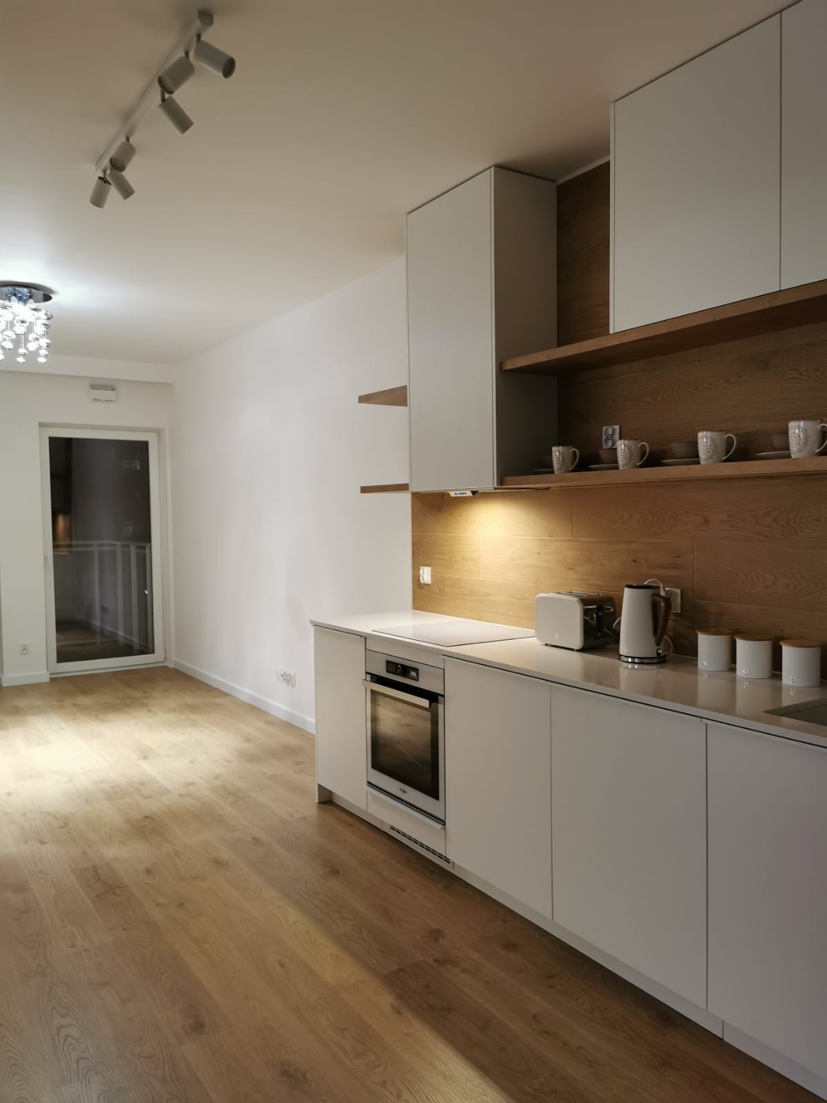 Nowoczesna kuchnia w minimalistycznym stylu z białymi frontami szafek i drewnianym panelem na ścianie, oświetlona punktowymi lampami sufitowymi i podświetleniem blatu, z widocznym sprzętem AGD...