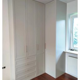 SN COMPANY SERHII NOVOMODNYI - Jasnoszara szafa narożna z czterema szufladami i długimi, smukłymi uchwytami, dopasowana do rogu pokoju z drewnianą podłogą i oknem.