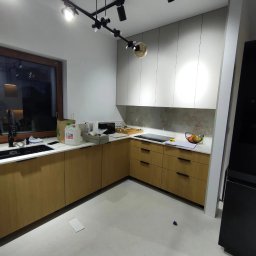 SN COMPANY SERHII NOVOMODNYI - Wnętrze kuchni w trakcie montażu: widoczne szafki dolne z drewnianymi frontami, białe szafki górne, blat z wyposażeniem, lampy sufitowe na szynie, okno i sprzęt AGD. Na podłodze leżą drobne elementy.