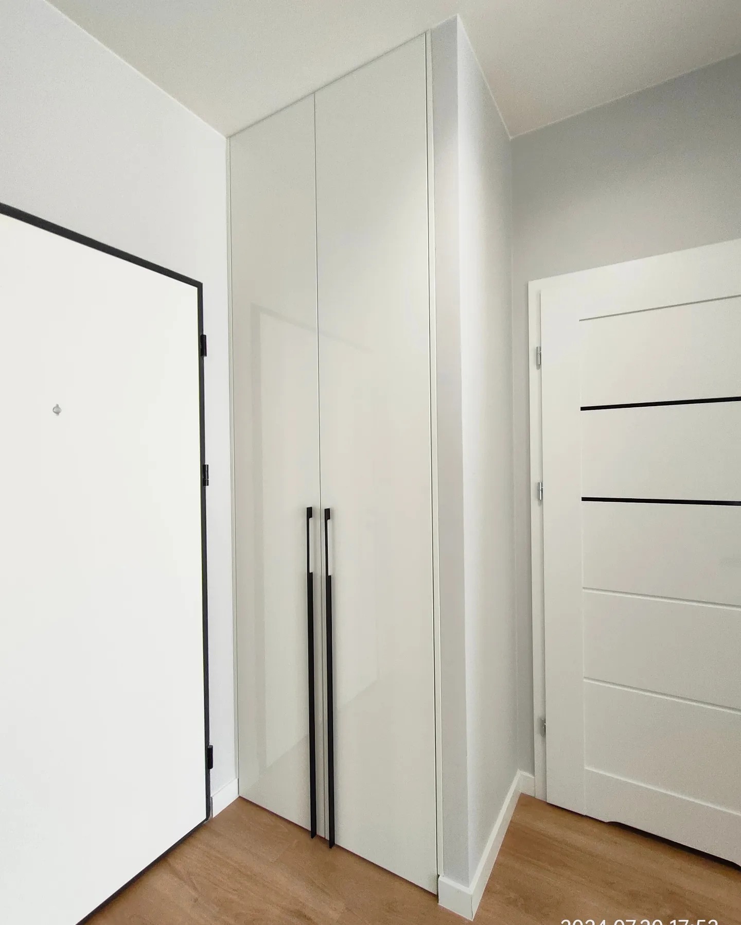 Biała szafa wnękowa z długimi, czarnymi uchwytami w przedpokoju, obok drzwi wejściowych z czarną ramą i drugich drzwi wewnętrznych.