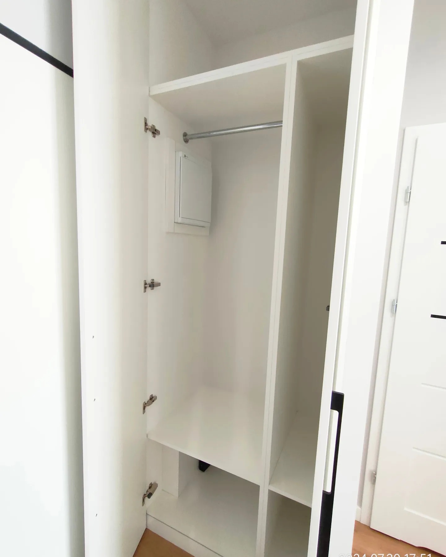 Wnętrze białej szafy z drzwiami na zawiasach, widoczna półka, drążek na ubrania i biała skrzynka elektryczna.